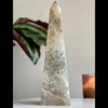 A polished agate tower sculpture with a geometric pattern, placed on a surface with a plant and a candle in the background.