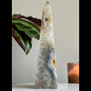 A polished agate tower sculpture with a geometric pattern, placed on a surface with a plant and a candle in the background.
