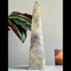 A polished agate tower sculpture with a geometric pattern, placed on a surface with a plant and a candle in the background.