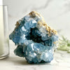 A large blue Celestite crystal geode on a marble surface with a candle and decorative greenery in the background.