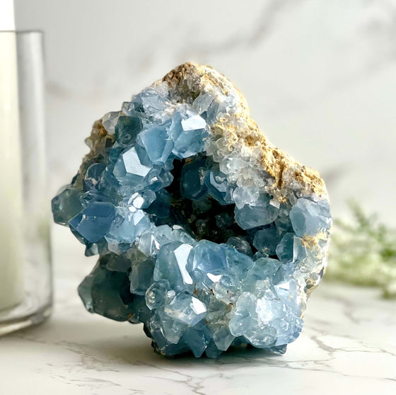 A large blue Celestite crystal geode on a marble surface with a candle and decorative greenery in the background.