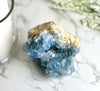 A large blue Celestite crystal geode on a marble surface with a candle and decorative greenery in the background.