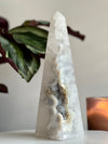 A conical sculpture made of druzy agate with a smooth, polished surface, placed on a white surface with a plant in the background.
