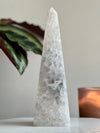 A conical sculpture made of druzy agate with a smooth, polished surface, placed on a white surface with a plant in the background.