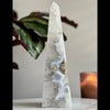 A conical sculpture made of druzy agate with a smooth, polished surface, placed on a white surface with a plant in the background.