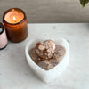 A polished agate heart-shaped stone on a marble surface with a candle and a plant in the background.