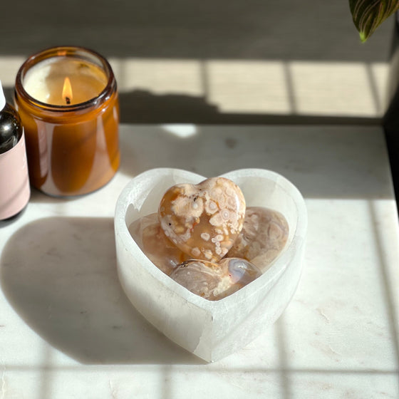 A polished agate heart-shaped stone on a marble surface with a candle and a plant in the background.