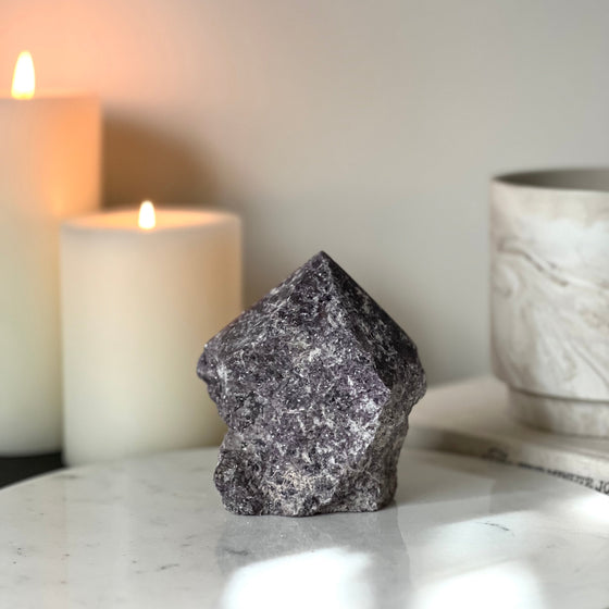 A semi-polished Lepidolite point crystal on a white marble stand with a background featuring candles and a book.
