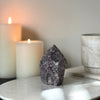 A semi-polished Lepidolite point crystal on a white marble stand with a background featuring candles and a book.
