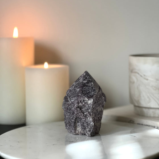 A semi-polished Lepidolite point crystal on a white marble stand with a background featuring candles and a book.
