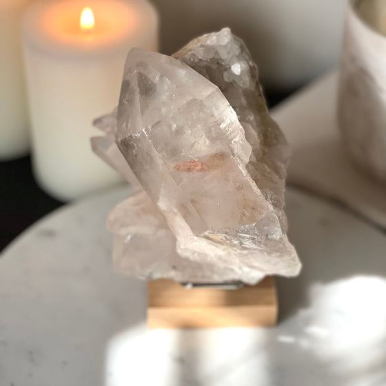 Pink Himalayan Samadhi Quartz on a wooden and silver stand with a blurred background featuring candles and a book.
