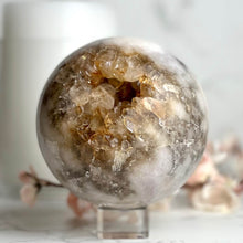  A polished sphere-shaped Pink Amethyst crystal with a clear center and golden-brownish periphery, placed on a clear stand against a marble background with some decorative flowers nearby.