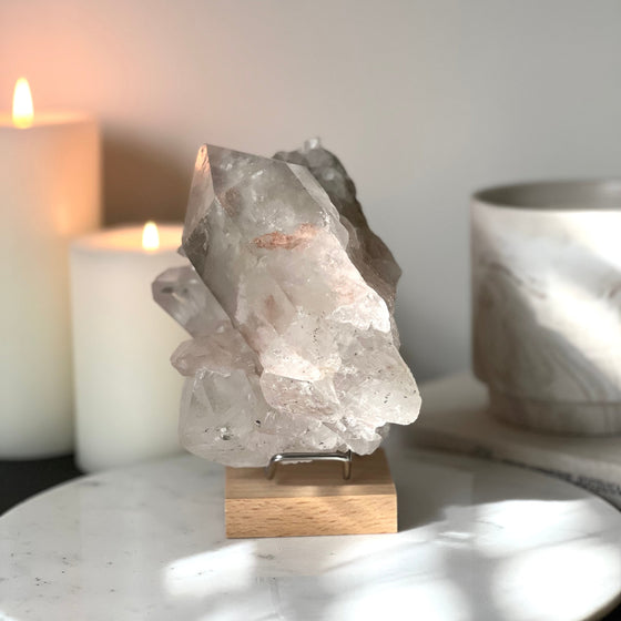 Pink Himalayan Samadhi Quartz on a wooden and silver stand with a blurred background featuring candles and a book.