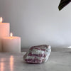A rough, pink tourmaline crystal on a marble surface with a candle and a green plant in the background.