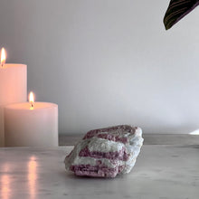  A rough, pink tourmaline crystal on a marble surface with a candle and a green plant in the background.