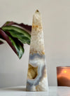 A polished agate tower sculpture with a geometric pattern, placed on a surface with a plant and a candle in the background.