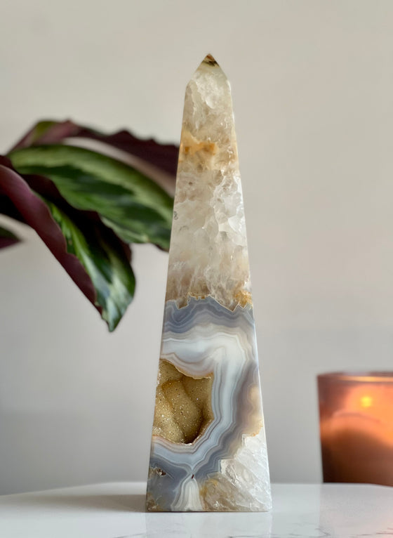 A polished agate tower sculpture with a geometric pattern, placed on a surface with a plant and a candle in the background.