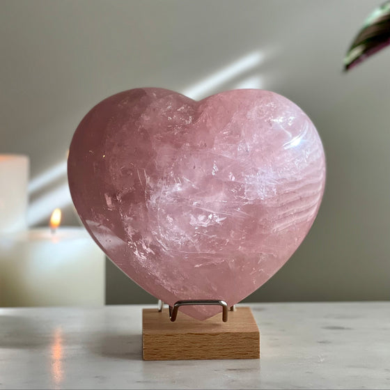 Large carved Rose Quartz heart on metal and wooden stand perched atop a marble base with a candle and plant in the background.