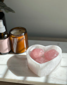  A polished, smooth, pink rose quartz heart-shaped palm stone, placed on a marble surface, with a shadow falling over the stone.
