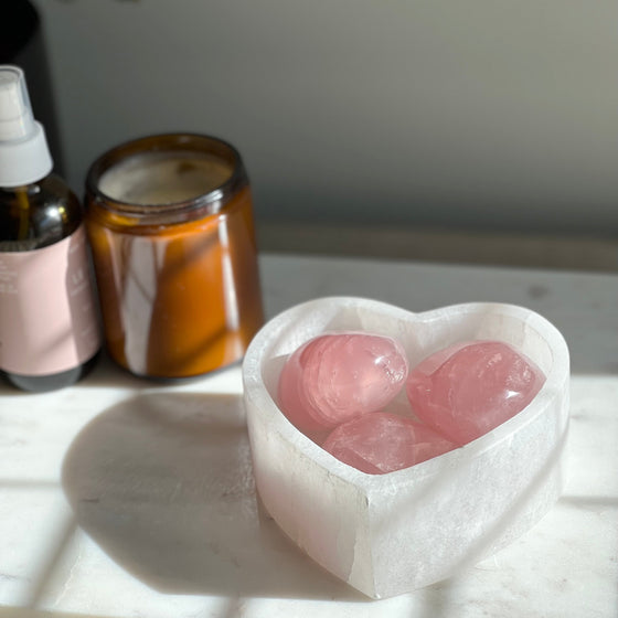 A polished, smooth, pink rose quartz heart-shaped palm stone, placed on a marble surface, with a shadow falling over the stone.